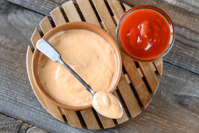 Yum Yum Sauce: The Irresistible Sauce That Will Make Your Taste Buds Dance – Discover How to Make It at Home!" 23 A single shot of a terracotta bowl filled with pink Yum Yum Sauce on a bamboo trivet, paired with a spoon and a small glass of ketchup on the side.