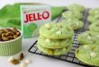 Pistachio Pudding Cookies: Easy Dessert Recipe 131 Stack of soft pistachio pudding cookies with white chocolate chips and chopped pistachios on a cooling rack, next to a box of pistachio pudding and a ramekin of whole pistachios