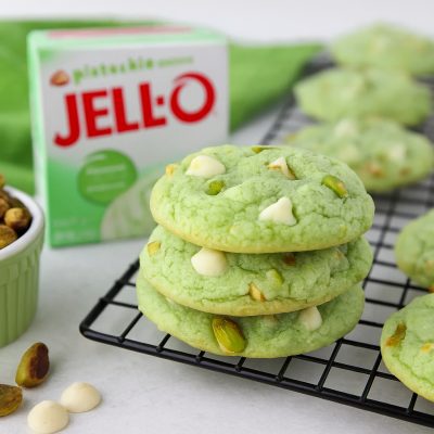 Pistachio Pudding Cookies: Easy Dessert Recipe 33 Stack of soft pistachio pudding cookies with white chocolate chips and chopped pistachios on a cooling rack, next to a box of pistachio pudding and a ramekin of whole pistachios